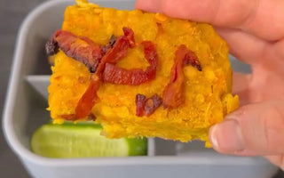Hand holding savory yellow baked dish with sun-dried tomatoes over bento box with cucumber slices