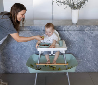 Baby in high chair with mess-free food catcher, mom feeding with smile